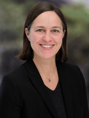 Photo of Nella Young smiling in front of a blurred natural background, wearing a black sweater and suit jacket